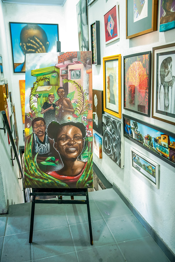 VAL_OPNI_GALERIAFAVELA_8.JPG Quadro em cavalete. Pintura mostra uma menina e três meninos negros sorrindo. Folhas rodeiam eles e há casinhas atrás