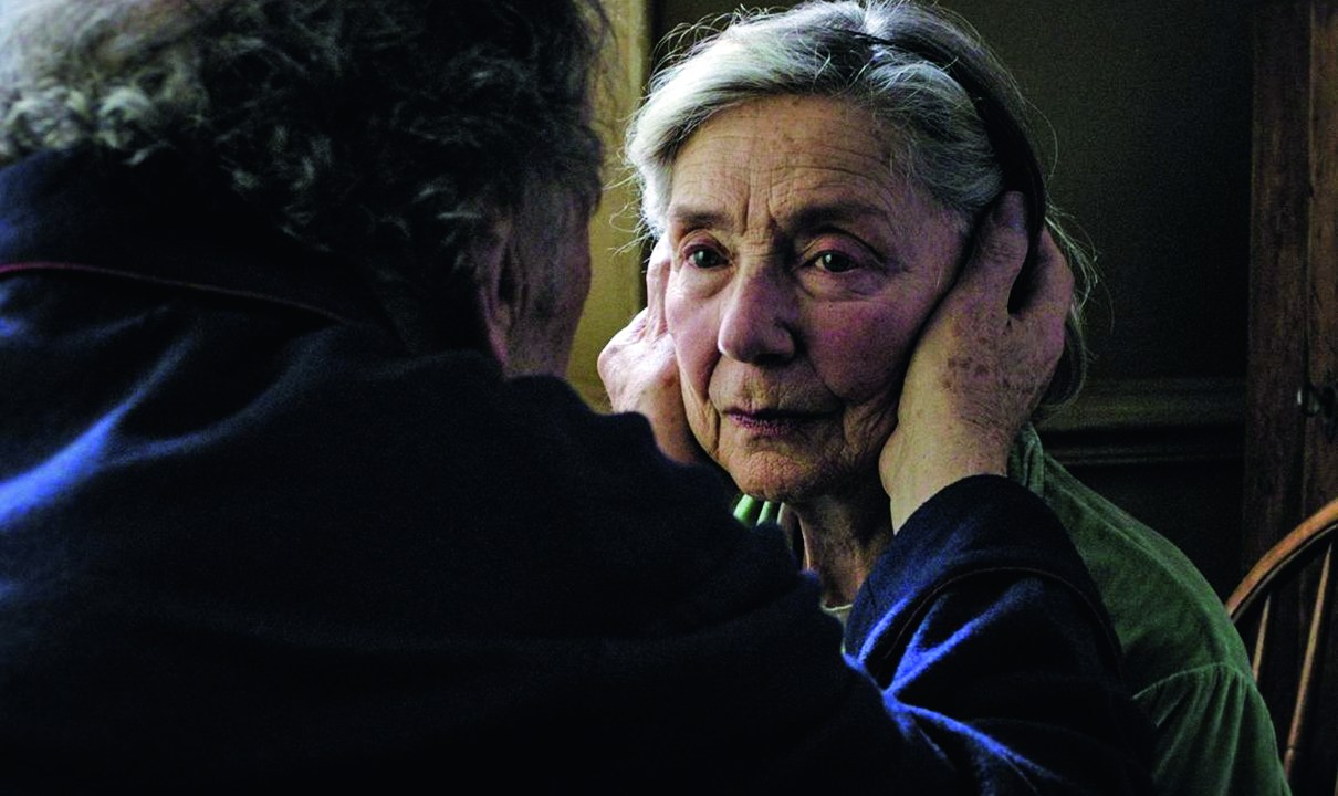 A imagem mostra um cena do filme Amor em que aparece o casal de velhinhos, um de frente para o outro, com o homem com as duas mãos no rosta da mulher