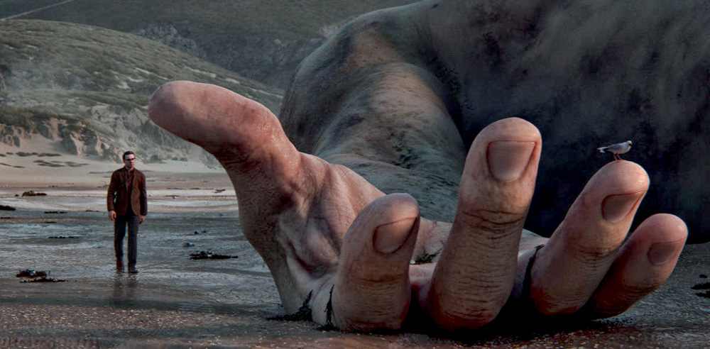 A imagem mostra uma pessoa ao lado de um mão gigantesca. Eles estão na beira de uma praia, próximo ao mar, com a mão já sobre a água.