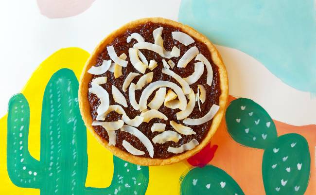 Foto aérea de torta de abóbora coberta por lascas de coco tostado sobre fundo colorido com desenho de cactus.