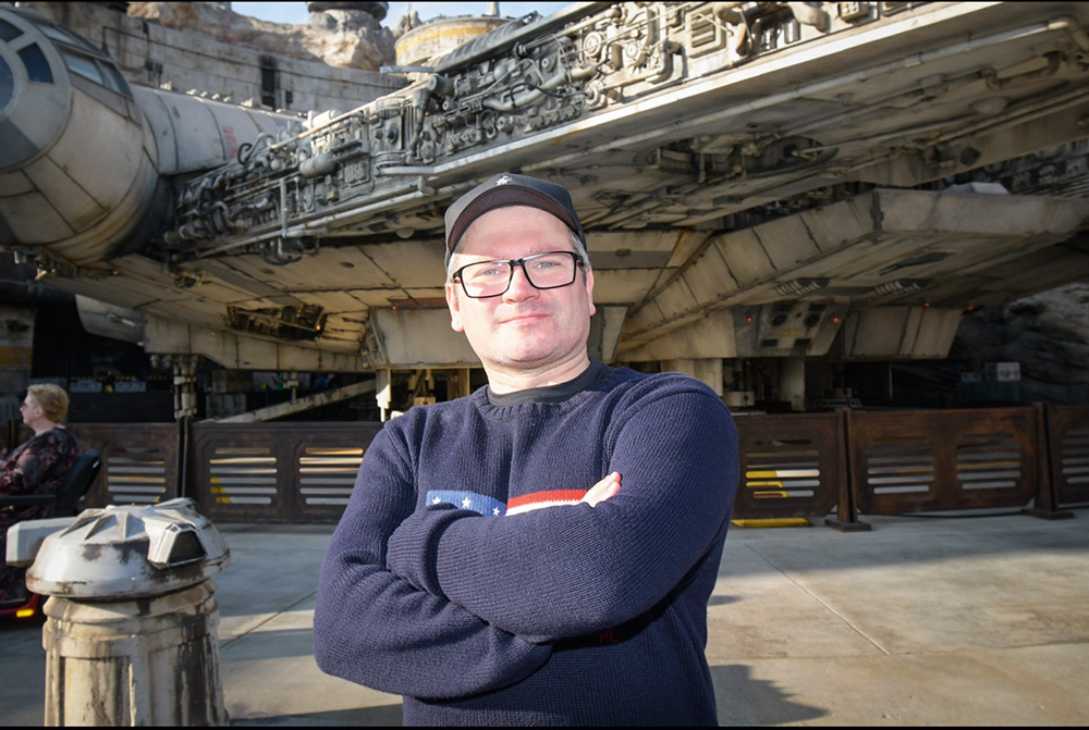 Carlos Henrique Marques posa de braços cruzados em frente a réplica de nave do Star Wars. Veste boné, óculos de grau e suéter de mangas longas na cor azul-escuro.