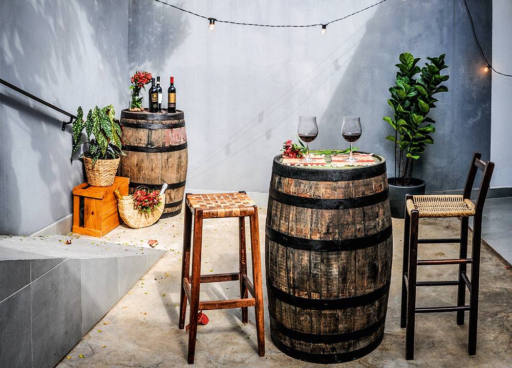 Quintal da Trattoria Mobile no Alto de Pinheiros, com dois barris um no canto esquerdo e outro à frente, em destaque, junto de duas banquetas de madeira com taças de vinho sobre ele. Corda de luzinhas passa pela parede azul ao fundo, com vazo de planta à direita.
