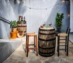 Quintal da Trattoria Mobile no Alto de Pinheiros, com dois barris um no canto esquerdo e outro à frente, em destaque, junto de duas banquetas de madeira com taças de vinho sobre ele. Corda de luzinhas passa pela parede azul ao fundo, com vazo de planta à direita.