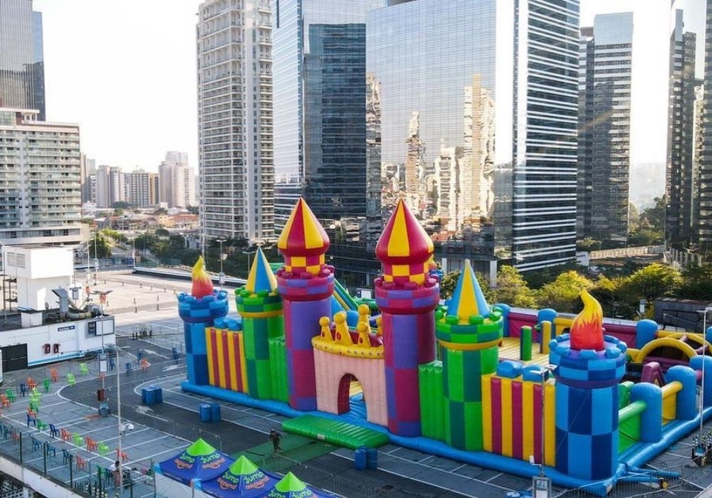 No estacionamento de um shopping, está uma estrutura inflável em formato de castelo colorida. Há prédios em volta