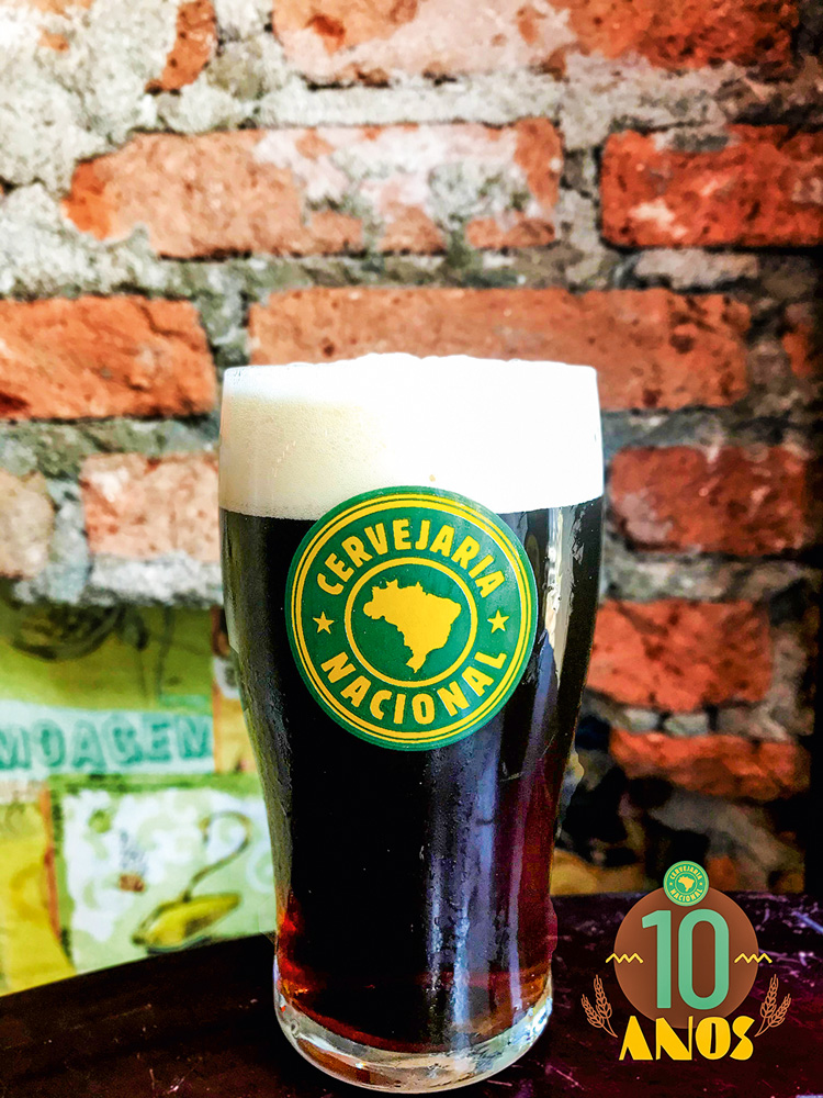 Drakes – Cervejaria Nacional Copo de chope com símbolo da Cervejaria Nacional completo com cerveja de coloração escura com colarinho.