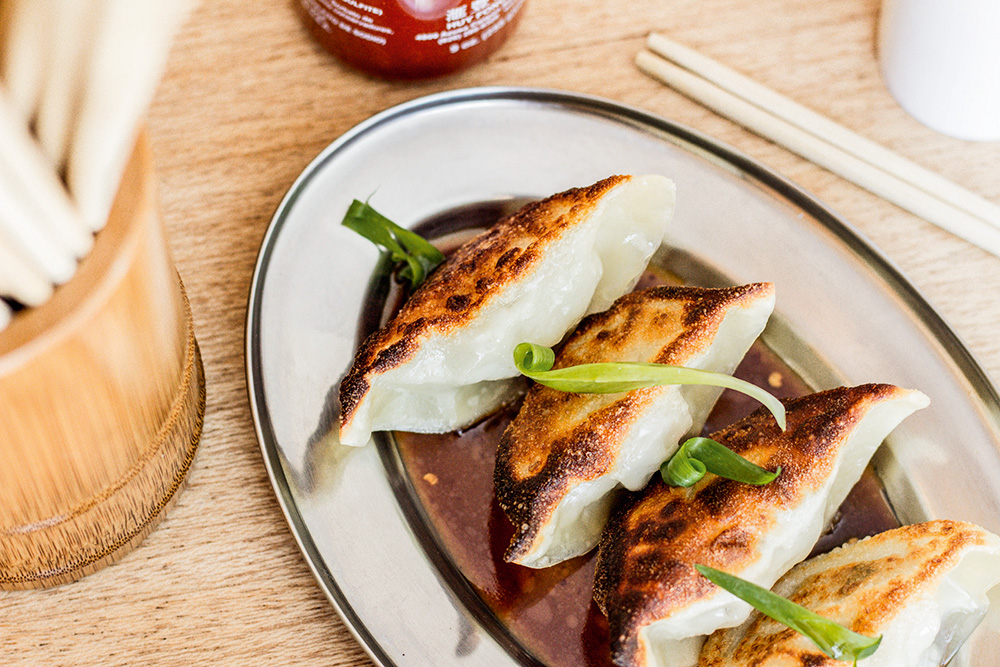 Foto tirada de cima de travessa de metal sobre mesa de madeira com quatro guiozas cobertos por folhas verdes.