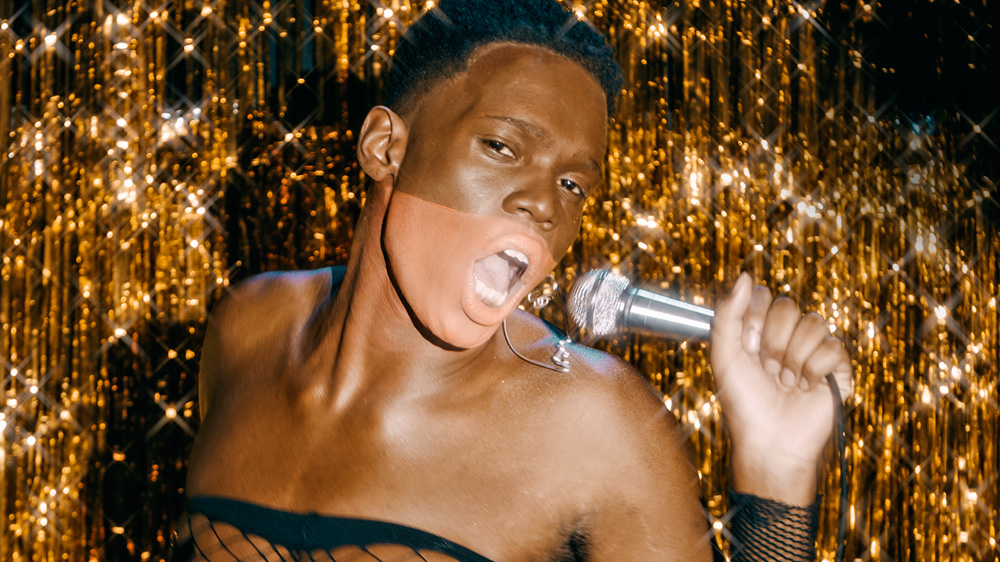 PASMA (REMIX) JUNIOR TOBAGO.jpg homem negro com detalhe no rosto cantando com uma cortina dourada brilhante no fundo