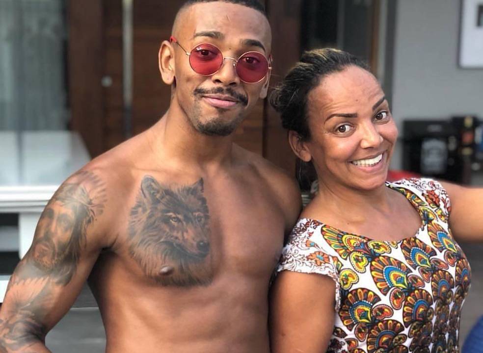 A imagem mostra Nego do Borel e sua mãe Roseli, ambos sorrindo para a foto.O cantor está de óculos vermelhos e sem camisa.