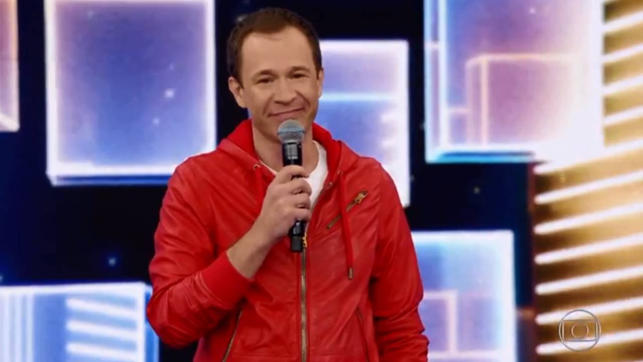 Na imagem mostra Tiago Leifert, com um microfone na mão, no cenário do Domingão do Faustão sorrindo