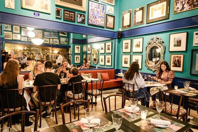 Salão do restaurante Ça-Va. Com paredes verde-água cobertas de quadrinhos e mesas com pessoas comendo em mesas nos cantos esquerdo e direito.