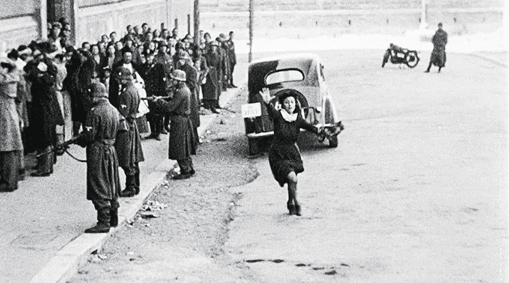 classicos5.jpg A imagem mostra uma rua cheia de pessoas, com militares e uma criança correndo no meio dela com o braço levantado