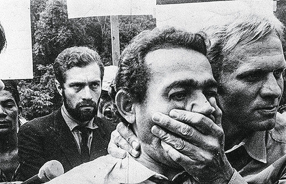 classicos7.jpg A imagem mostra um homem em meio a alguma aglomeração, com outro homem de seu lado colocando a mão sobre sua boca, como se tentasse tampá-la