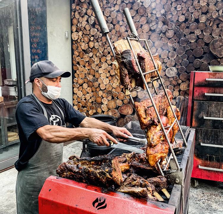 Churraqueiro de máscara cuidando de carnes em espetos eque estão sobre a grelha de churrasqueira vermelha.