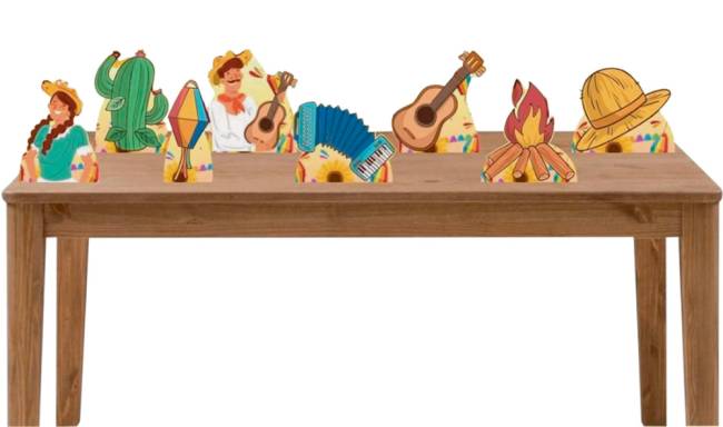 Uma mesa de madeira tem displays de desenhos de festa junina. Balão, fogueira, violão, chapéu de palha, cacto e homem e mulher