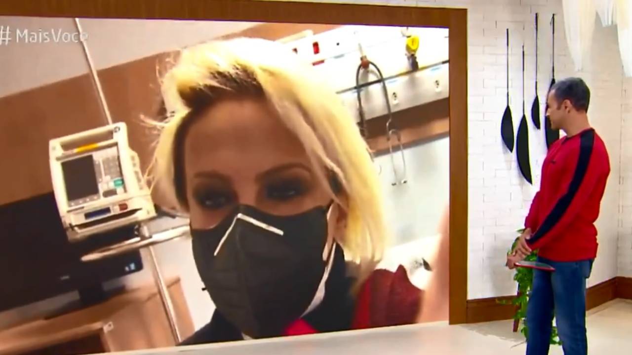 A imagem mostra o apresentador Fabrício falando com Ana Maria através de um telão no programa Mais Você. Ela está de máscara, em um quarto de hospital, ele está de pé no cenário olhando para a tela.
