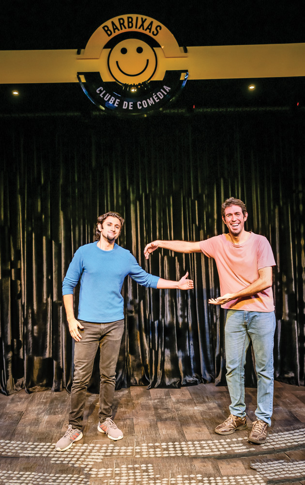 Clube Barbixas de Comédia Dois homens fazem pose de abraço, mas com distanciamento social, em um palco de um clube de comédia. Eles estão sorrindo