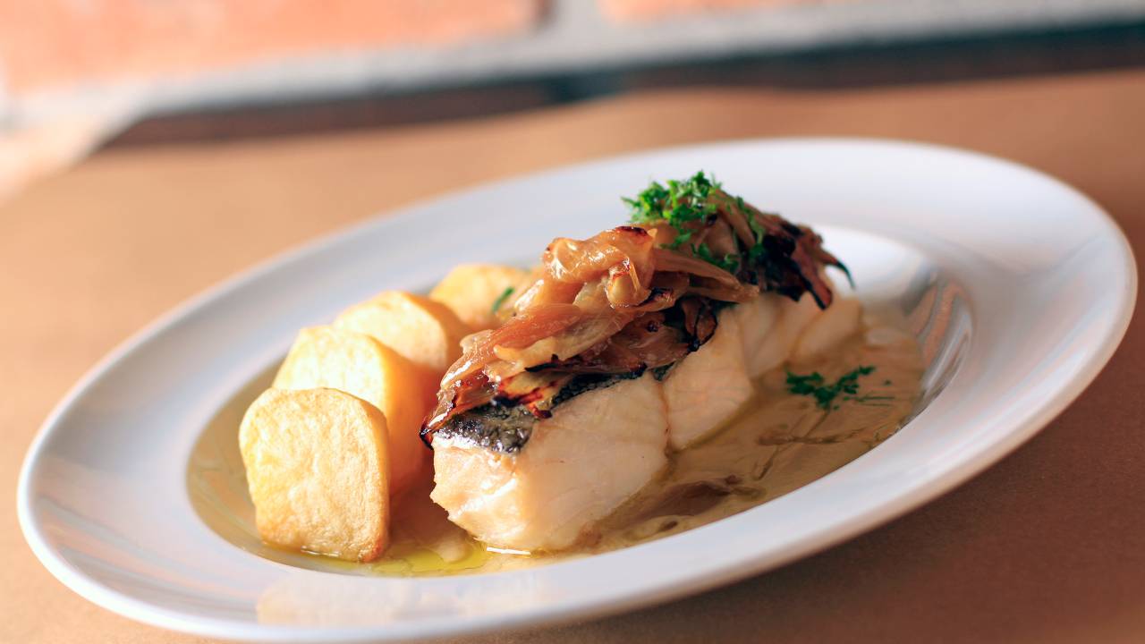 Prato oval com posta de bacalhau coberto por cebola ao lado de batatas.