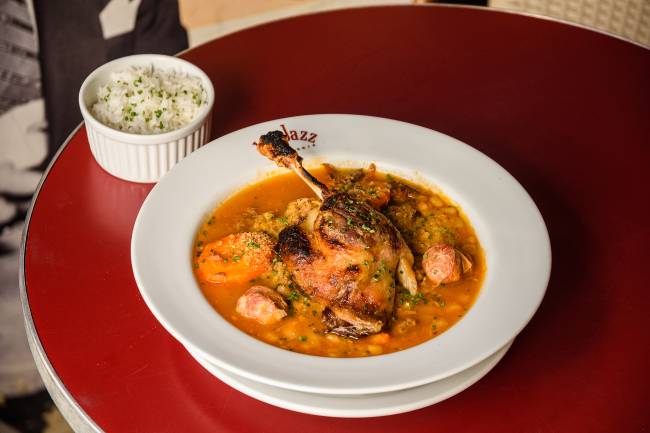 Cassoulet com coxa de frango assado sobre o cozido no prato fundo do Le Jazz sobre mesa vermelha ao lado de potinho de arroz.