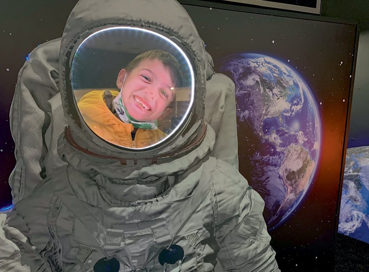 Menino dentro de uma estrutura que imita uma roupa de astronauta sorri e olha para a câmera