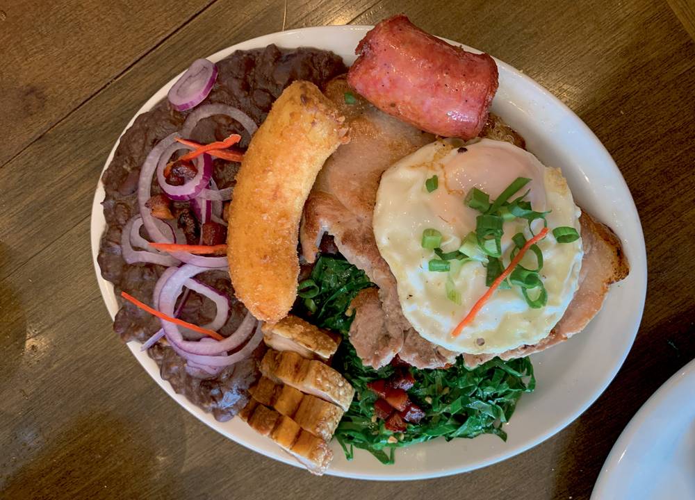 Prato oval de louça branca sobre mesa de madeira com virado à paulista (tutu de feijão coberto por cebola, banana frita, torresmo, linguiça, ovo frito, bisteca e couve).