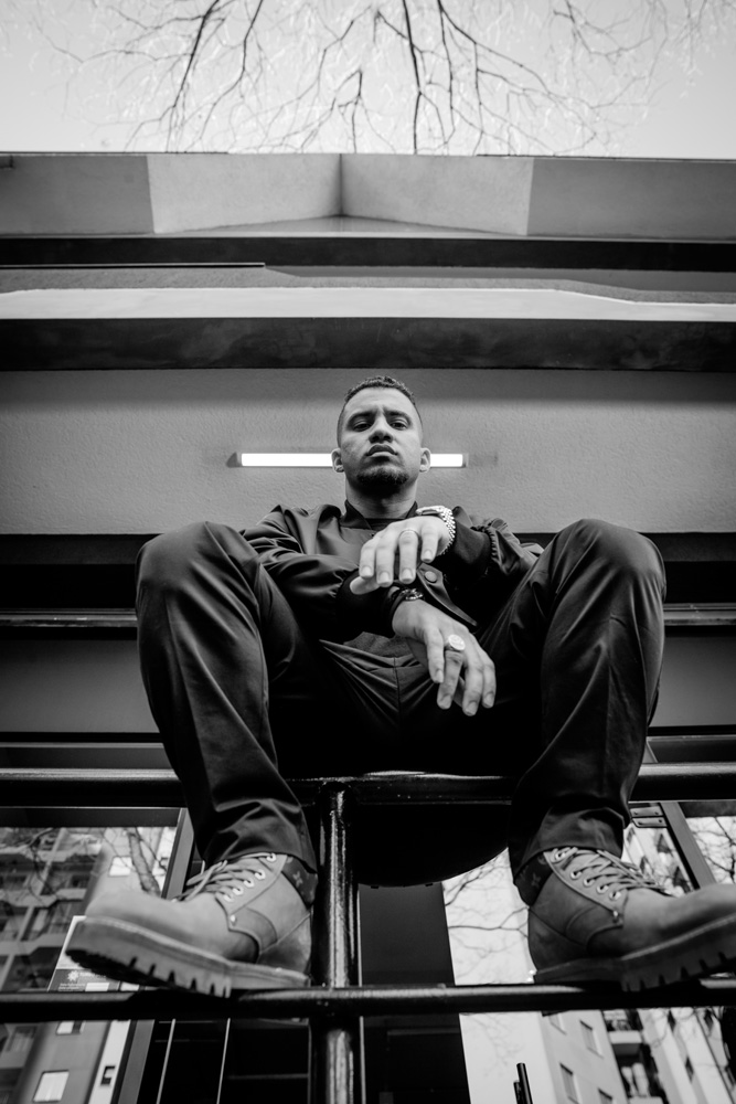 KondZilla-Konrad-Dantas A imagem mostra Konrad sentado em cercadinho de metal bem em frente a sua produtora. A foto é de um ângulo inferior, vendo primeiro os pés dele apoiados na grade e o rosto mais distante.