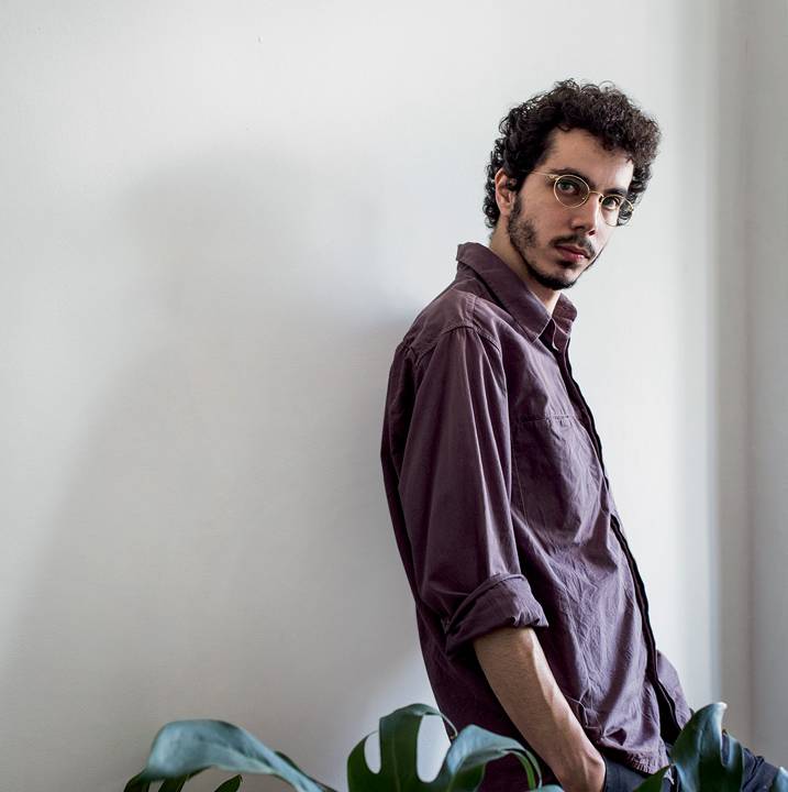 Luan Cardoso encara a câmera de óculos e camisa de cor escura. Aparece encostado na parede. Em primeiro plano, a foto exibe uma planta da espécie costela-de-Adão