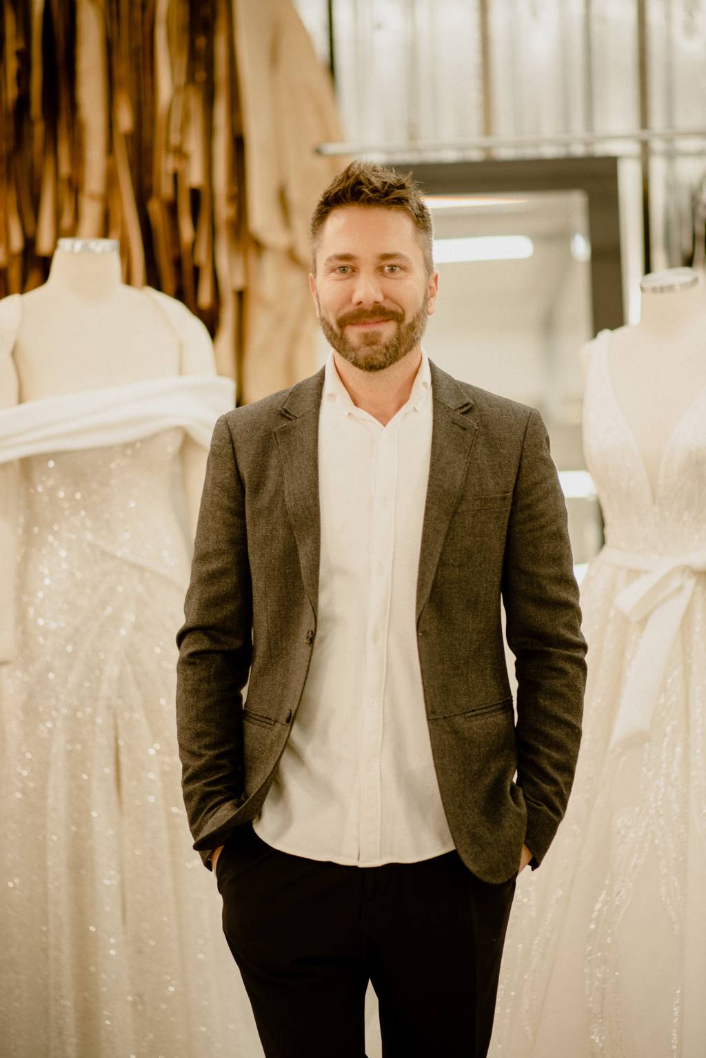 Lucas Anderi – Disney Princess Wedding Collection (Jeff Porto) (1) Lucas Anderi posa com as mãos no bolso vestindo paletó escuro e camisa branca. Dois vestidos de noiva aparecem de fundo.