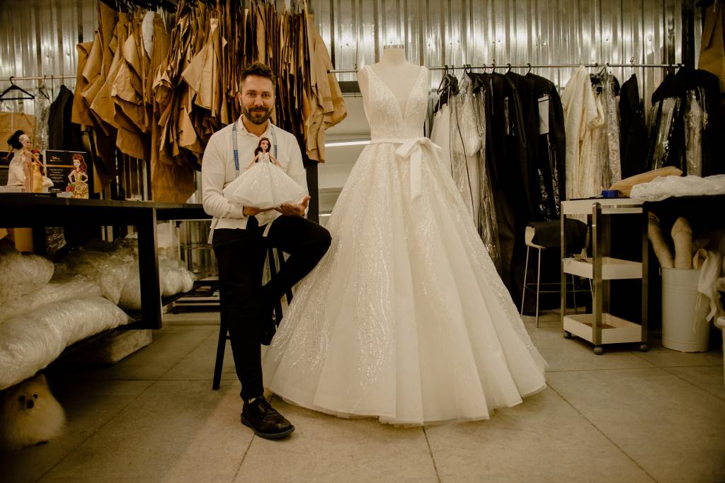 Lucas Anderi – Disney Princess Wedding Collection (Jeff Porto) (4) Lucas Anderi aparece sentado ao lado de vestido de noiva. Veste calça preta e camisa branca, com fita métrica no pescoço e segurando boneca.