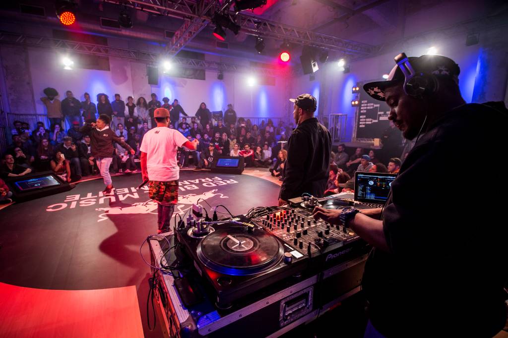 Participantes de Red Bull Francamente, evento de rap (Fabio Piva-Red Bull Content Pool Foto mostra palco escuro com holofotes na parte de cima. No meio, dois rappers batalham com rimas e poesias faladas. Ao fundo, uma plateia assiste o evento. Em primeiro plano, um DJ aparece controlando sua mesa de mixagem.