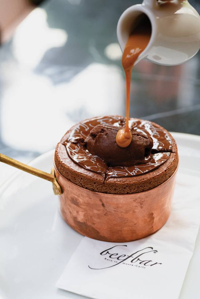 Beefbar Suflê de chocolate em panelinha de cobre sobre prato de louça branca do Beefbar recebendo calda de caramelo de jarrinha de louça branca.