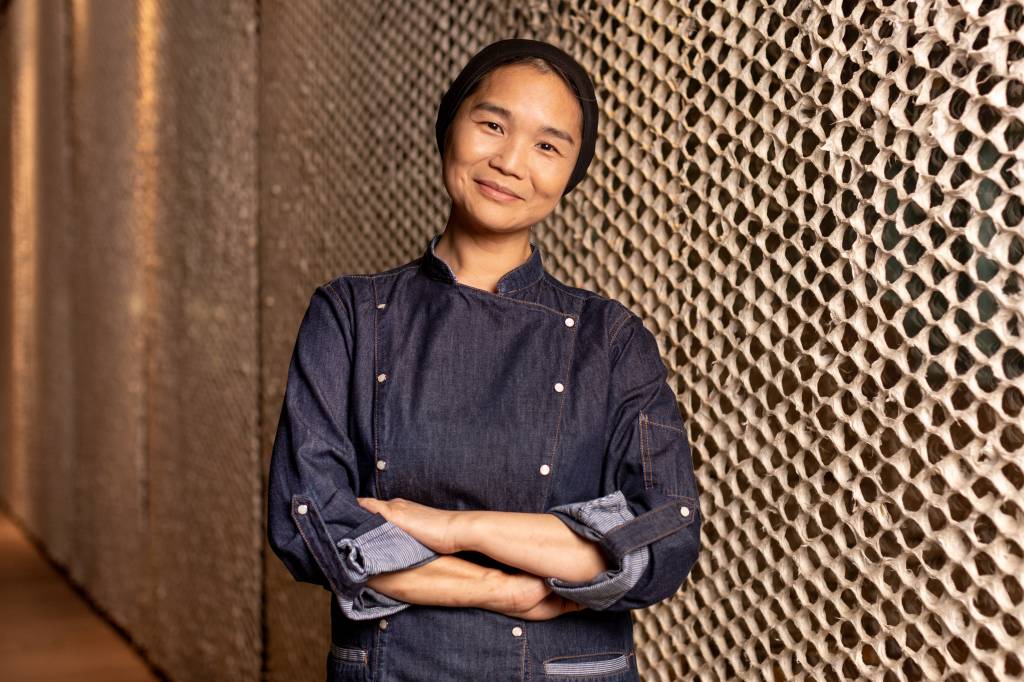 Telma Shiraishi A chef Telma Shiraishi sorrindo de braços cruzados em frente à parede de textura rendada.