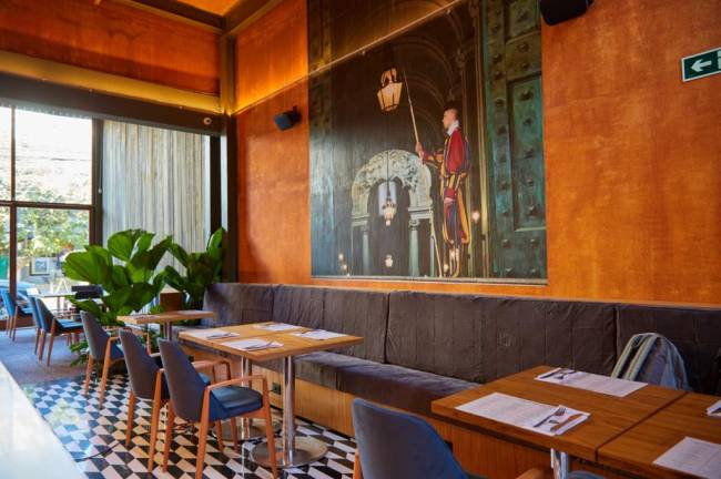 Modern Mamma Osteria Salão do restaurante Moma Modern Mamma Osteria em Pinheiros. Com chão quadriculado preto e branco, paredes alaranjadas e mesas à direita.