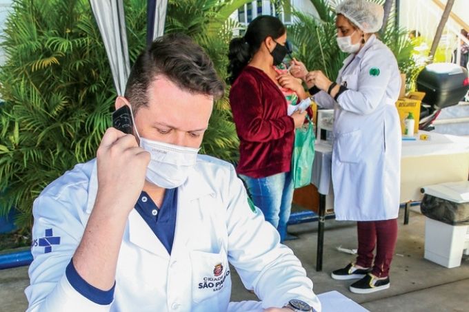 Danilo Mesquita em primeiro plano ao celular utilizando máscara e de avental da cidade de são paulo. ao fundo, uma pessoa sendo imunizada por uma enfermeira