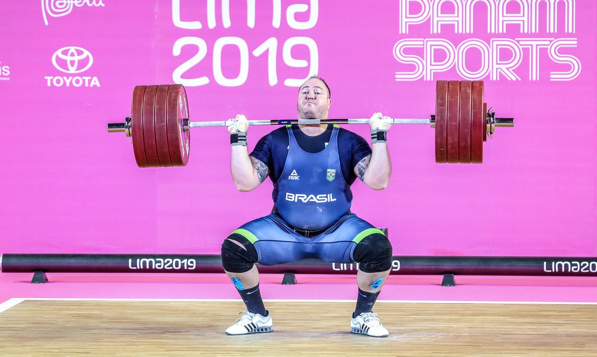 Imagem, com fundo rosa, mostra Fernando Reis levantando peso durante Pan-americano de Lima, no Peru, em 2019
