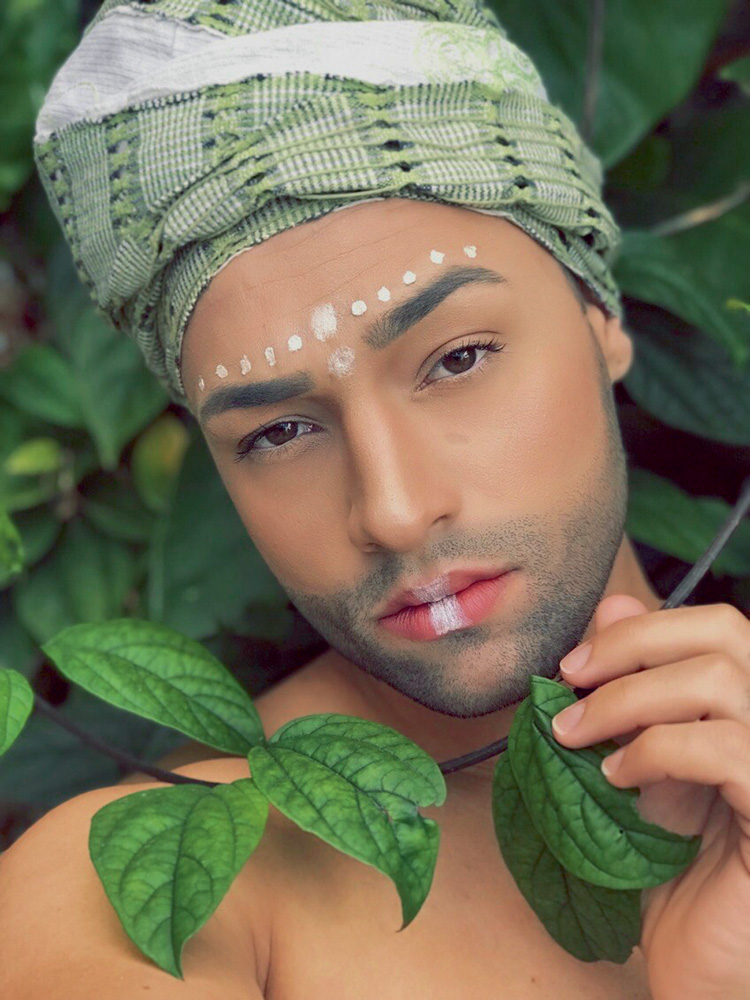 Jean Fernandes – influenciadores TikTok Homem está apoiado em algumas folhas verdes, usando turbante e com algumas pinturas brancas no rosto