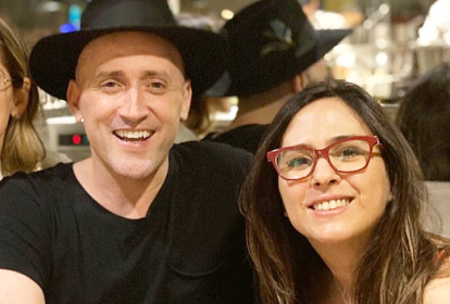 Imagem mostra Paulo Gustavo e Tatá Werneck abraçados, sorrindo. Tatá usa óculos de grau e Paulo, um chapéu