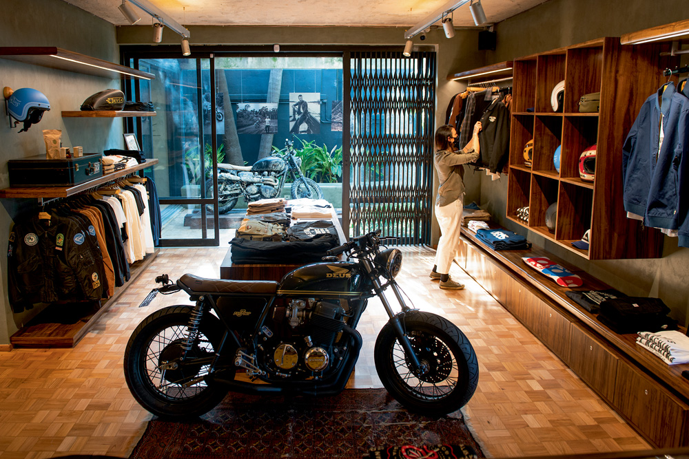 Deus Ex Machina A imagem mostra a fachada da Deus Ex Machina. Nela, há roupas em exibição e uma moto no centro da loja.