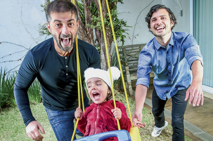 A imagem mostra Rogerio e Gustavo empurrando a filha Alice em um balanço. Os três estão sorrindo.