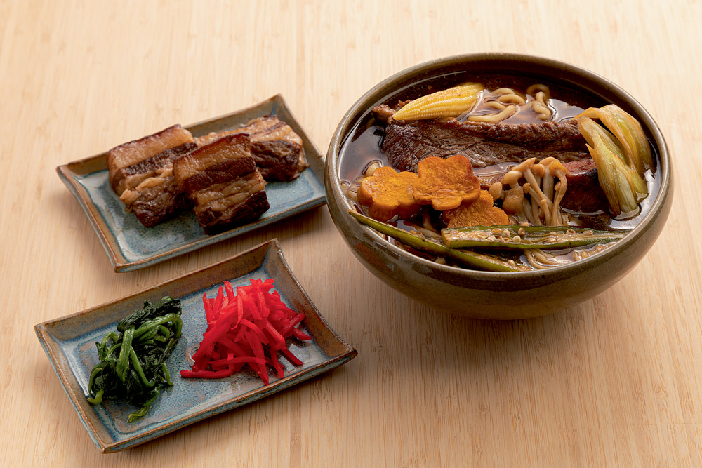 Sobre mesa de madeira, lámen do Jojo Ramen ao lado de bandejinhas de cerâmica com ingredientes para ser preparado em casa.