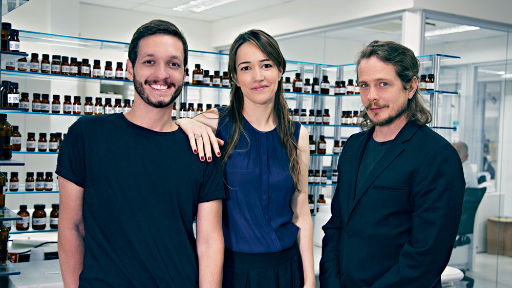 Isaac-Fany-Fábio-L’Envie-perfumistas-rotina-fragrâncias-perfumes Isaac, Fany e Fábio sorrindo para a foto entre prateleiras de frascos de fragrâncias
