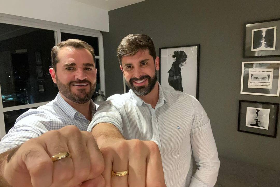 A imagem mostra Marcelo e Frankel, vestidos de branco e mostrando os punhos fechados para a câmera. É possível ver entre os dedos as alianças deles.