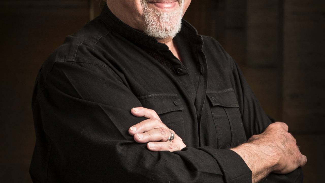 A imagem mostra Paulo Coelho, de braços cruzados, em um fundo escuro e de pé.