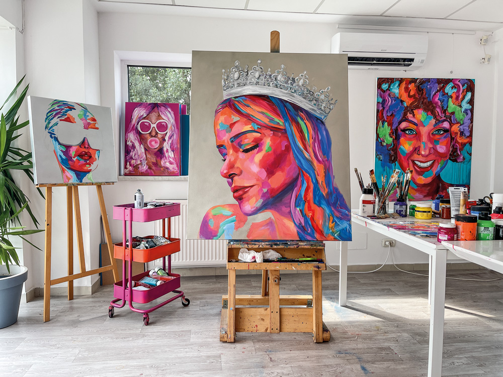 Retratos Natalia Martins (Ramona Pintea) Obra de Ramona Pintea aparece erguida em um ateliê. O quadro exibe o rosto de Natalia Martins em versão colorida (rosa, azul e roxo) com uma coroa na cabeça).