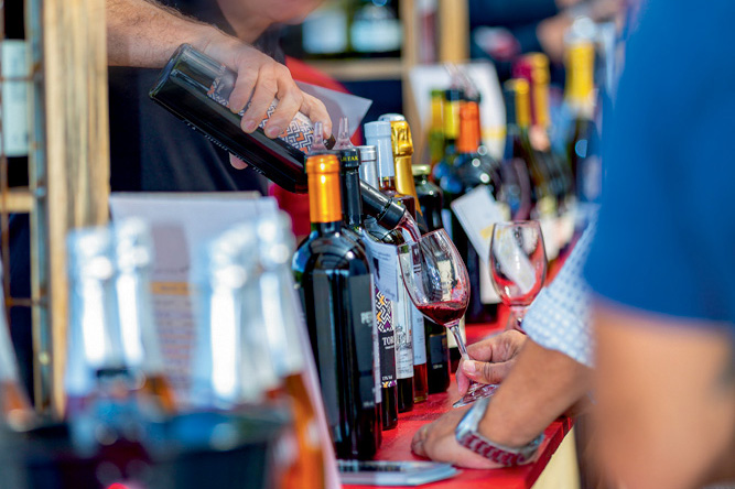 Garrafas de vinho enfileiras expostas em estande. Uma mão serve vinho tinto em uma taça.
