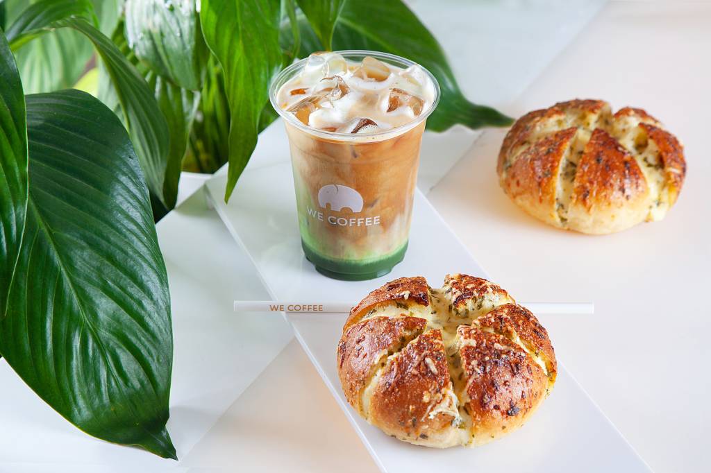 Achados ELO – We Coffee Pão de alho arredondado sobre bancada branca e café com doce de leite e matchá num copo de plástico transparente atrás.