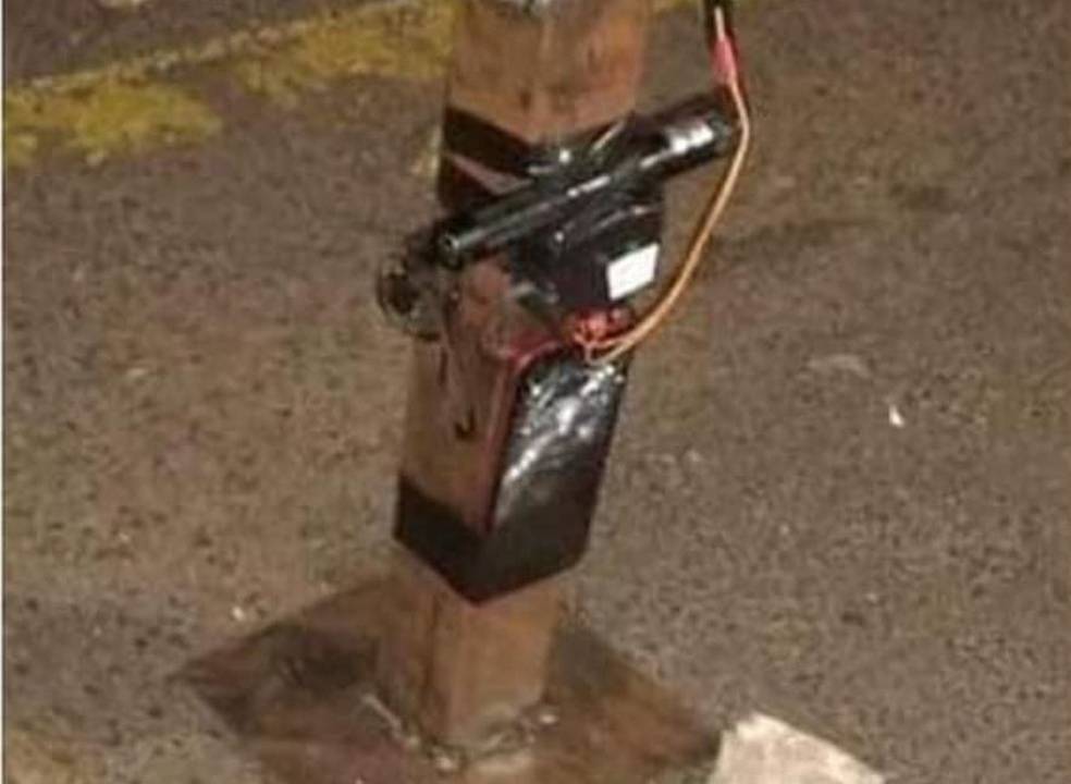 Imagem mostra pedestal no meio de rua, com fios e placa grudadas por meio de fita isolante no item de metal