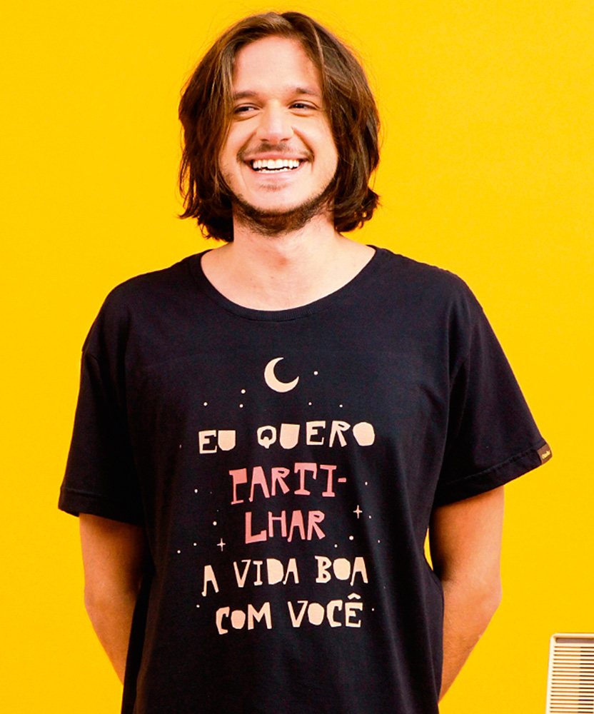 camiseta 2.png Um homem sorri em um fundo amarelo vestindo uma camiseta preta com a frase