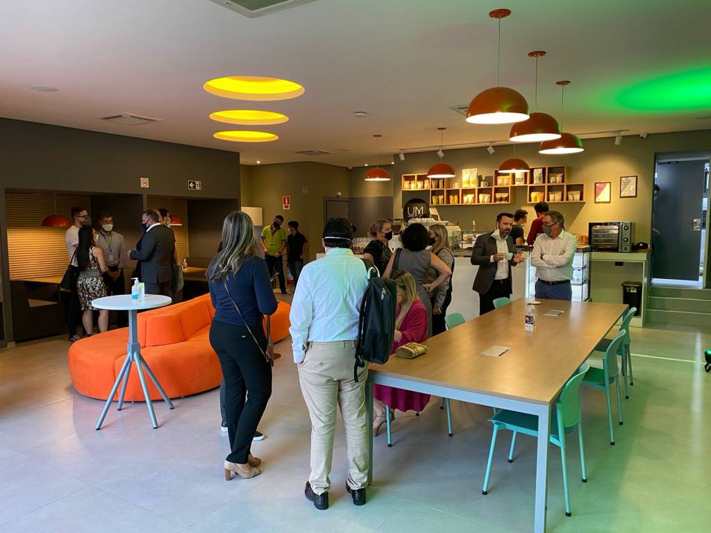 Um Coffee Co. Salão da unidade da Faria Lima do Um Coffee Co. com pessoas em pé, cabines de mesas à esquerda e uma mesa retangular comprida à direita.