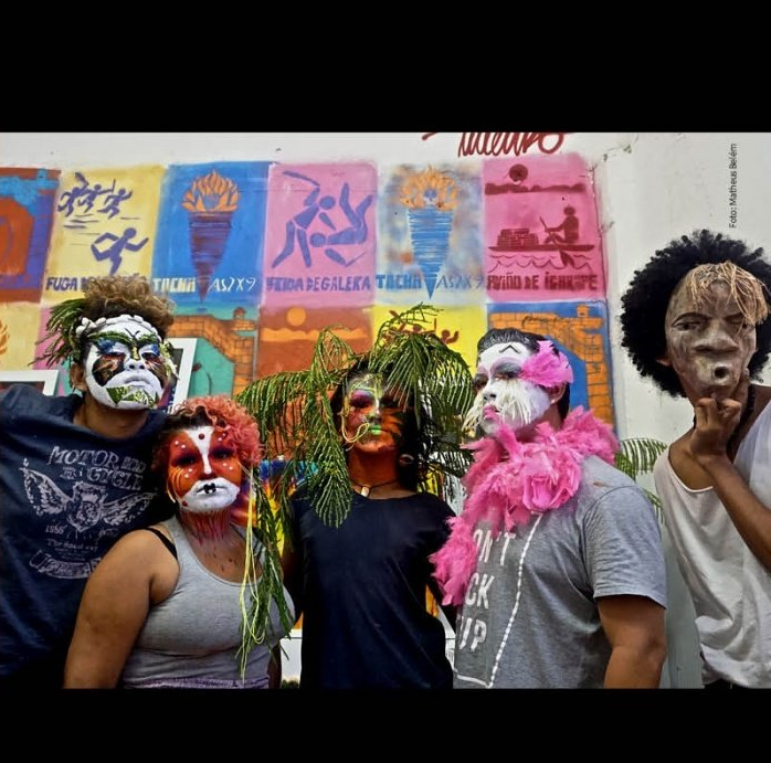 Pessoas com máscaras e maquiagem, no fundo um painel com cartazes