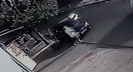 Imagem mostra frame de vídeo de segurança em que carro aparece encostado perto de calçada; próximo ao veículo, mulher sai de perto do carro com criança no colo
